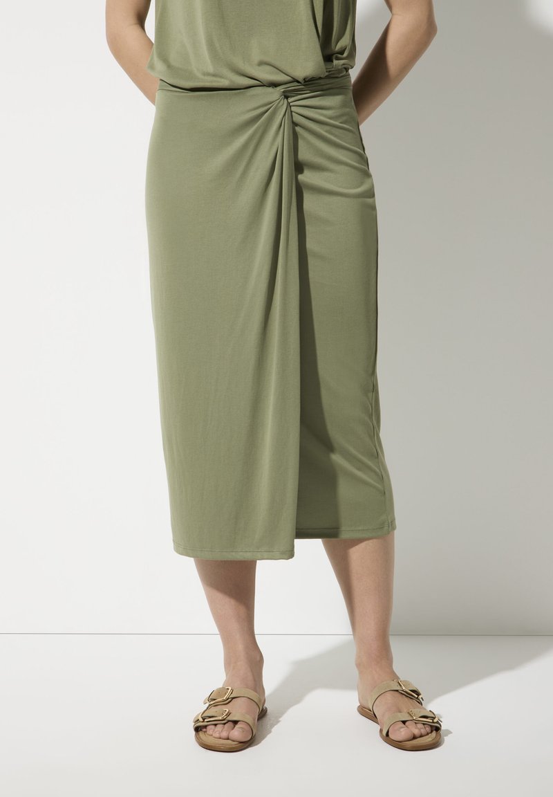 Olivegrüner Midirock aus weichem Stoff mit einem verdrehten Detail an der Taille und einer geraden Silhouette; kombiniert mit hellbraunen Sandalen.
