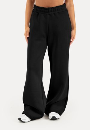 Person iført løse sorte bukser med elastisk talje og hvide sneakers, stående mod en ensartet lys baggrund.