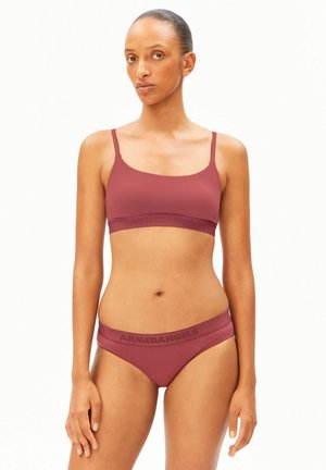 Femme aux cheveux attachés, portant un ensemble de lingerie mauve assorti avec le logo "ARMEDANGELS", debout devant un fond blanc uni.