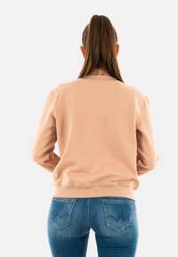 Maglione girocollo rosa chiaro con colletto e polsini a coste, indossato sopra jeans di denim blu scuro con tasche posteriori angolate.