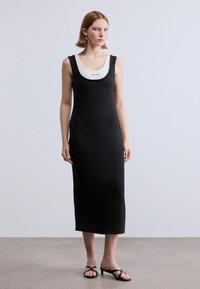 Μαύρο εφαρμοστό φόρεμα με στρογγυλή λαιμόκοψη και λευκή λεπτομέρεια στο γιακά, κατασκευασμένο από απαλό ύφασμα. Το μοντέλο φοράει μαύρα σανδάλια με τακούνι.
