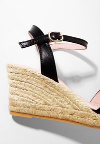 Sandale compensée à bride de cheville en cuir noir avec plateforme en jute tressé et boucle dorée sur fond blanc.
