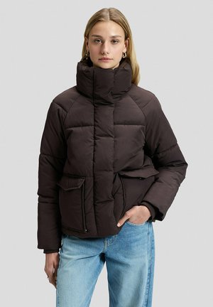 Frau mit langen blonden Haaren trägt eine dunkle Steppjacke mit hohem Kragen und blaue Jeans, stehend vor einem einfarbig hellgrauen Hintergrund.