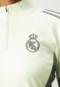 Gai zaļš sporta žakets ar augstu apkakli, pelēkiem akcentiem un lielu pelēku Real Madrid logo uz krūtīm. Viegla tekstūra.