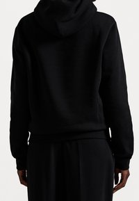 Schwarzer Hoodie mit weicher, strukturierter Oberfläche. Verfügt über eine Kapuze und gerippte Bündchen. Trägt eine passende schwarze Hose und ist von hinten zu sehen.
