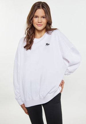 Ung kvinde med brunt hår iført en løs hvid sweatshirt med teksten "Mrs." på brystet og sorte bukser, stående med hånden på hoften.