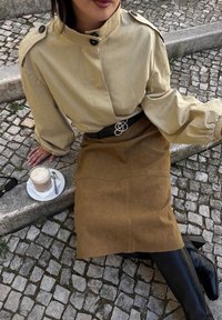 Bežinė palaidinė su peties mygtukais, ruda zomšinė sijonas, juodas diržas su sidabrinėmis detalėmis, juodos kelių ilgio botai, šalia lėkštėje kava.