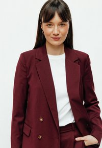 Blazer bordeaux à double boutonnage avec des boutons dorés, associé à un t-shirt blanc à col ras du cou. Tissu lisse, design structuré, avec poches latérales.