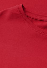 Maglietta rossa con una texture liscia, scollo rotondo e maniche corte. Il tessuto sembra elastico, con un colore uniforme e dettagli di cucitura visibili.