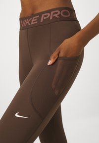 Leggings sportivi marroni con una ampia vita di un marrone scuro con la scritta "NIKE PRO" in rosa chiaro. Include una tasca laterale e una texture liscia.