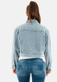Chaqueta de mezclilla azul clara con corte corto, adornada con tachuelas plateadas en los hombros y las mangas, usada sobre una blusa blanca, combinada con jeans azules.