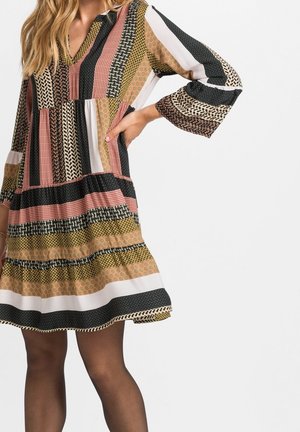 Femme portant une robe multicolore à motifs avec des manches longues et des collants noirs transparents, debout avec une main sur la hanche.
