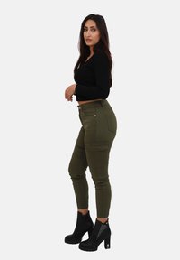 Crop top noir à manches longues associé à un pantalon cargo vert olive avec des poches latérales. Femme portant des bottines à talons noires avec des clous.