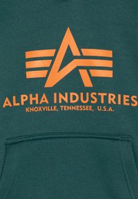 Logo di Alpha Industries con una "A" stilizzata e tre strisce orizzontali sopra il testo che legge "Alpha Industries Knoxville, Tennessee, U.S.A." su tessuto verde.