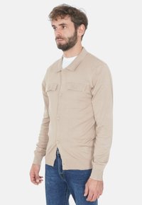 Cardigan beige con colletto, due tasche sul petto e chiusura con bottoni, realizzato in un materiale morbido, indossato sopra una t-shirt bianca e jeans blu.