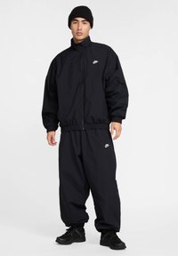 Nike svart treningsdress med en glidelåsjakke med elastiske mansjetter og snøring i midjen, kombinert med løse, innsnevrede bukser og svarte joggesko.