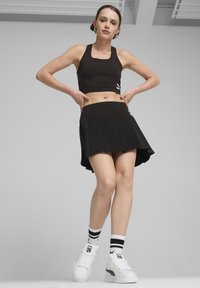 Fekete sportmelltartó, fekete pliszírozott szoknyával párosítva. A modellen fehér sportcipők vannak zöld kiegészítőkkel és fekete-fehér csíkos zoknikkal.