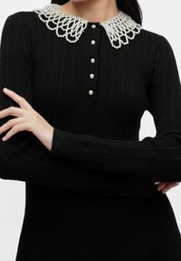 Pull côtelé noir à manches longues, patte de boutons en perles et col blanc orné de perles, bras croisés sur le torse.