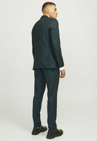 Tmavozelený strihový oblek s vyrezávanými klopami, slim fit a textúrovanou tkaninou. V kombinácii s čiernymi topánkami, z pohľadu zozadu.