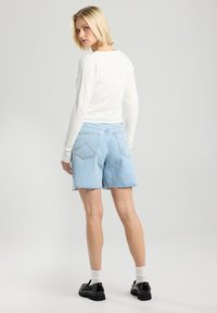 Valkoinen kiero pitkähiha toppi, yhdistettynä vaaleansinisiin farkkushortseihin, joissa on risat reunat ja takataskut. Mustat kengät ja valkoiset sukat.