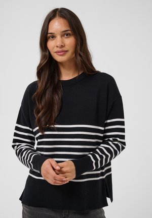 Femme aux longs cheveux bruns portant un pull noir à rayures horizontales blanches et un pantalon foncé, debout devant un fond uni.