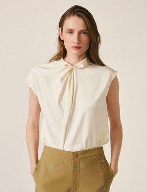 Femme aux cheveux longs portant un chemisier sans manches crème avec un col noué et un pantalon vert olive orné de motifs floraux brodés.