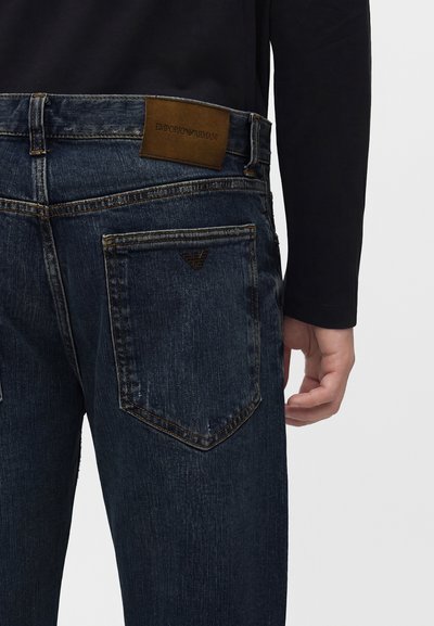 Σκούρο μπλε τζιν από denim με καφέ δερμάτινη ετικέτα Emporio Armani στη μέση και μεταλλικό λογότυπο στην πίσω τσέπη, φορεμένο με μαύρο μακρυμάνικο τοπ.