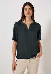 Blusa a maniche corte in tessuto verde scuro, con scollatura a V, texture morbida e dettagli arricciati sulle spalle, indossata con pantaloni beige chiaro.