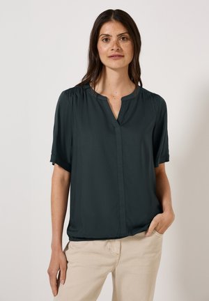 Blouse à manches courtes en tissu vert foncé, avec un col en V, texture douce et détail froncé aux épaules, portée avec un pantalon beige clair.
