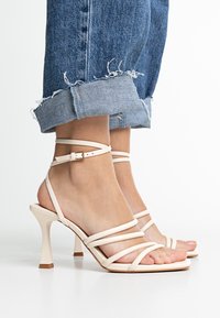 Sandali con tacco alto e cinturini in morbido materiale crema con punta quadrata, dotati di cinghie alla caviglia e design con tacco a blocco. Indossati con jeans risvoltati.