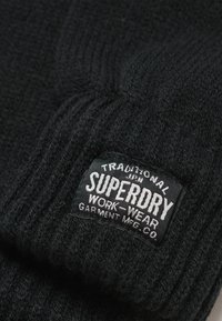 Gorro de punto negro con textura acanalada y un parche rectangular etiquetado "SUPERDRY WORK-WEAR GARMENT MFG. CO." en texto blanco.