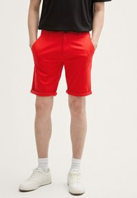 Pantalones cortos rojos hechos de una tela ligera, con un frente plano, bolsillos inclinados y dobladillos con vuelta. Combinados con zapatillas blancas y calcetines.
