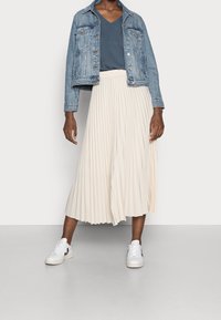 Jupe midi plissée beige clair associée à un haut bleu à col en V et une veste en jean délav ée. Des baskets blanches avec des accents noirs complètent le look.