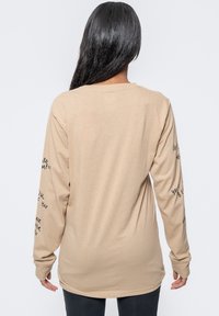 Long-sleeve beige t-shirt with text printed on sleeves, featuring a relaxed fit and soft, lightweight fabric. Hemline curves slightly.