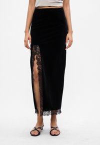 Fekete bársony maxi szoknya, magas hasítékkal és bonyolult csipkeborítással. Sima tapintás, testhezálló dizájn és elegáns sziluett.
