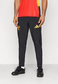 Muškarac nosi crne fudbalske trenirke Belgije s žutim Adidas logotipom i crne fudbalske kopačke na jednobojnoj pozadini.