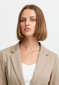 Beiger Blazer mit strukturiertem Design, Reverskragen und glatter Oberfläche, getragen über einem weißen Oberteil. Der Hintergrund ist schlicht weiß.