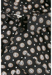Chemise en tissu noir avec des motifs circulaires blancs et marron et de petits points blancs, avec des boutons noirs et un col et des poignets pliés.