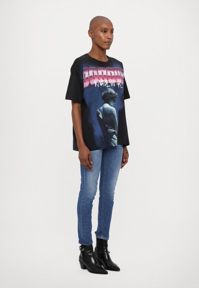 Camiseta negra oversized con un estampado gráfico que presenta una estatua y un texto en degradado de rosa a azul. Llevada con unos vaqueros azules y botas negras de tobillo.