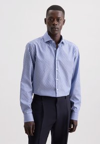 Uomo che indossa una camicia a righe verticali blu e bianche, infilata in pantaloni neri, in piedi contro uno sfondo chiaro e semplice.