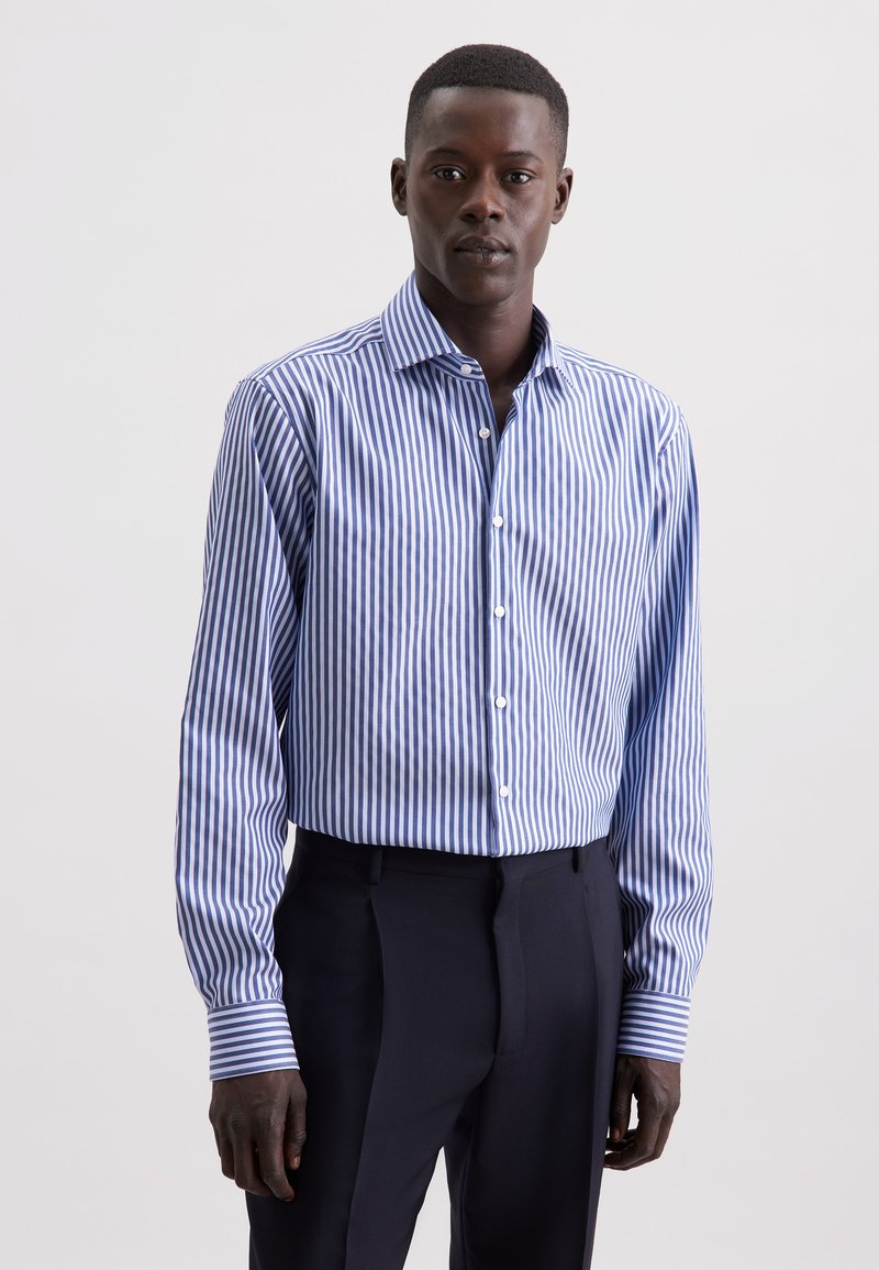 Uomo che indossa una camicia a righe verticali blu e bianche, infilata in pantaloni neri, in piedi contro uno sfondo chiaro e semplice.