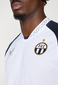 Tricou sportiv alb cu accente bleumarin, cu un design în formă de V, țesătură texturată, logo auriu cu dungi negre și cusături decorative la guler.