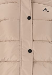 Beige pufferjakke med en quiltet tekstur, der har sorte knapper og et lille sort logo på den øverste højre side.