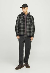 Zwart-wit geruite flanellen shirt over een grijze hoodie, gecombineerd met zwarte broek en bruine veterschoenen. Een zwarte pet maakt de outfit compleet.