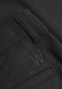 Juodas audinys su tekstūruota paviršiumi, su siuvinėta kišenės detale ir iškilniu "W" logotipu. Minimalistinis dizainas ir monochrominė spalvų schema.