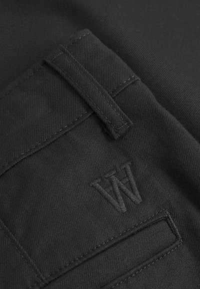 Tissu noir avec une surface texturée, comportant un détail de poche cousue et un logo "W" en relief. Design minimaliste et palette de couleurs monochromes.