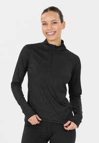 Smilende kvinde iført en sort langærmet zip-up træningsbluse og matchende leggings mod en ensfarvet hvid baggrund.