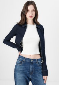 Marineblå denimjakke med krage, glidelås og langermet, over en hvit kort topp og lyseblå høy midje jeans.