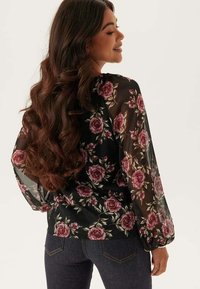 Svart blommig blus med långa, genomskinliga ärmar, prydd med rosa rosor och en avslappnad passform, tillsammans med mörka denimjeans.