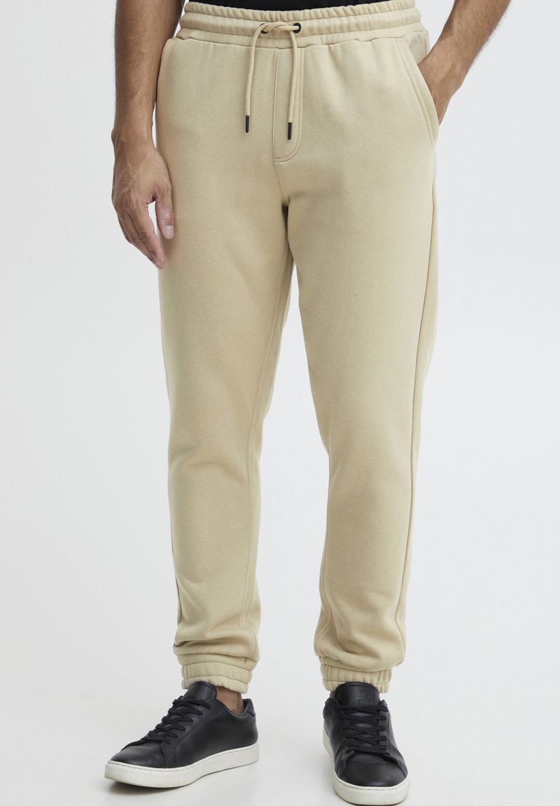 Beige Sweatpants mit elastischem Bund und Bündchen, ausgestattet mit Seitentaschen und verstellbarem Kordelzug, kombiniert mit schwarzen Sneakers.
